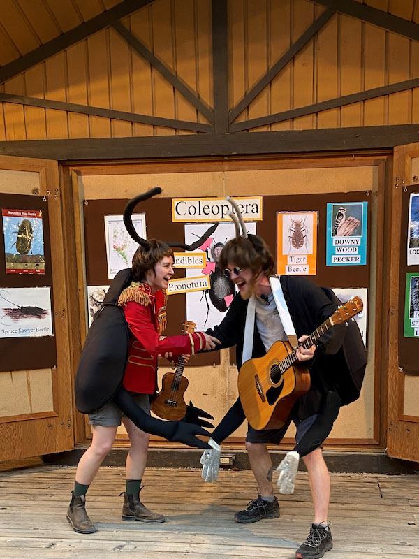 Two interpreters dancing. Both are dressed as beetles, with antennae and multiple arms. One is wearing round glasses, playing a guitar, and has a haircut like The Beatles. The other holds a ukulele.