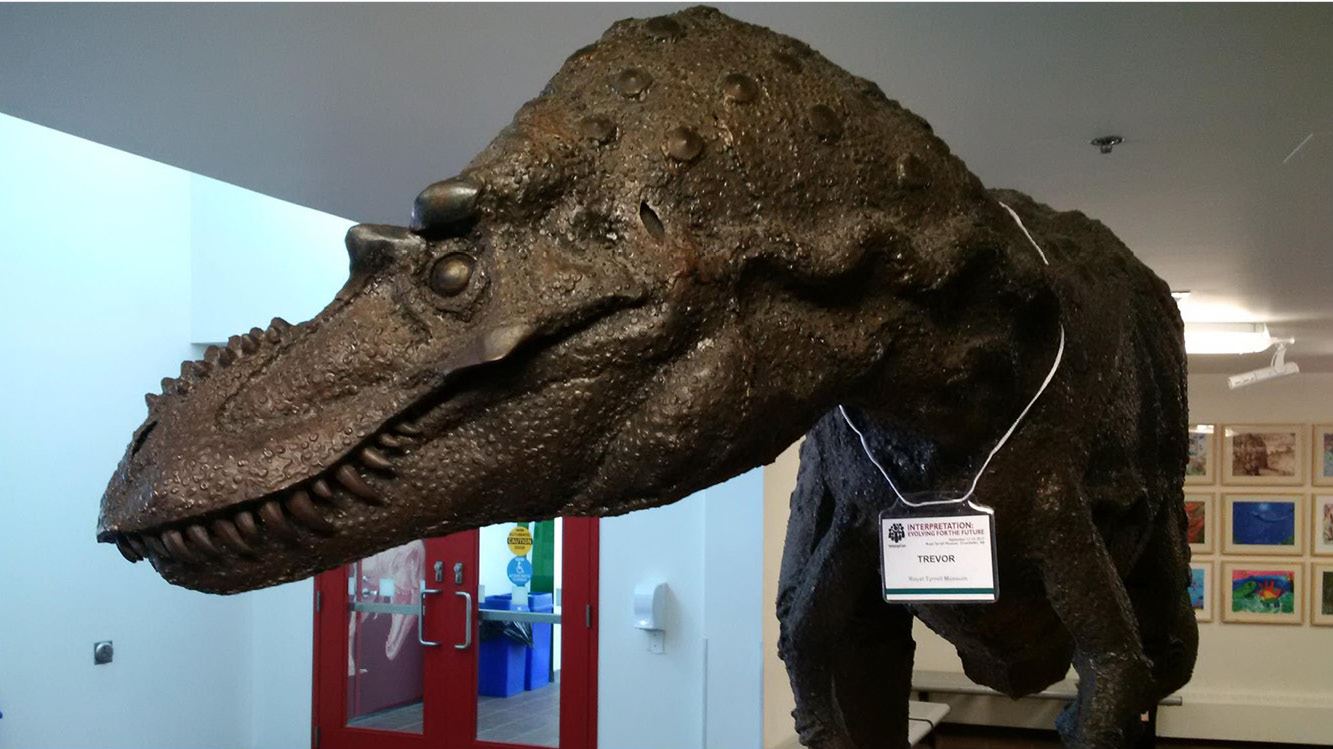 A life-sized statue of a dinosaur wears an Interpretation Canada nametag around its neck. The nametag reads “Trevor.”